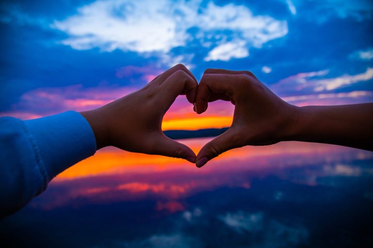 Zwei Hände von zwei Menschen formen ein Herz vor einem bunt leuchtenden Himmel bei Sonnenuntergang