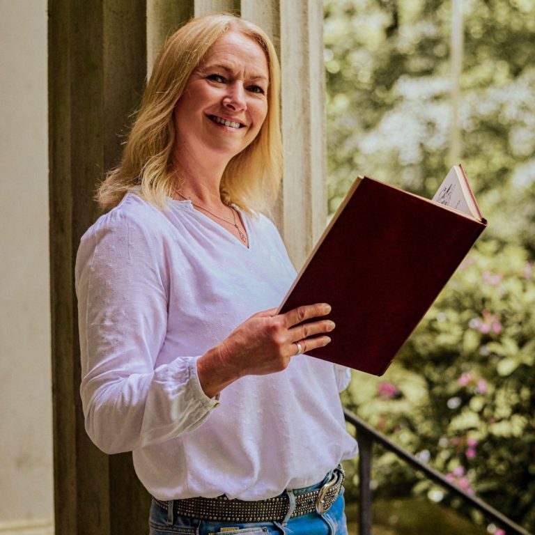 Blonde Frau, weiße Bluse lehnt draußen an Säule mit Buch in der Hand