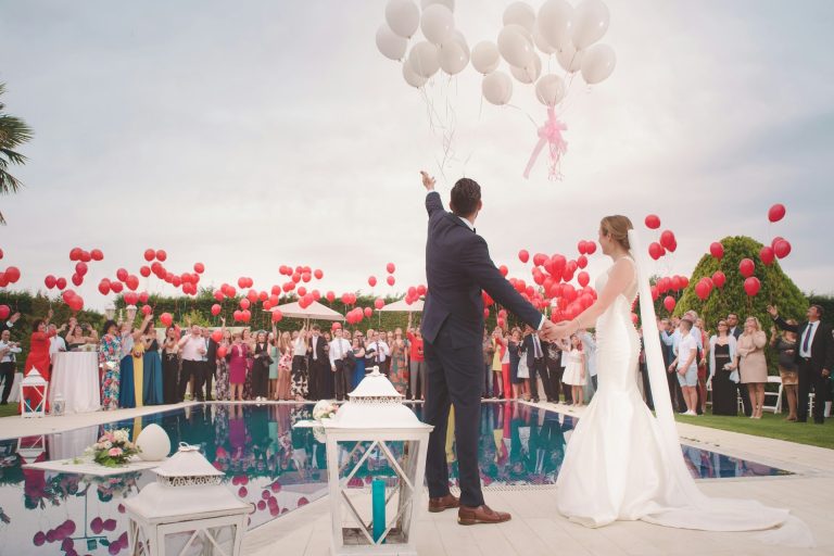 Ein Brautpaar steht mit seinen Gästen um einen Pool und lässt Luftballons steigen